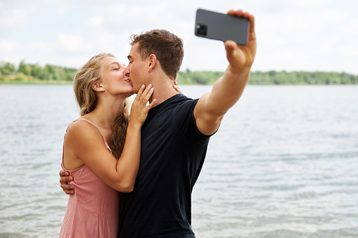 a young man takes a selfie with his girlfriend while they walk on the beach by a lake