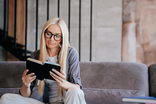Puzzled beautiful blonde Caucasian young woman in glasses reading book with bored face expression sits on cozy couch at home. Gorgeous Scandinavian businesswoman checking agenda.