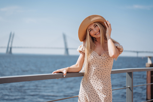 Cheerful Swedish beautiful young woman in beige dress standing at embankment of  bay in hat against sea and huge bridge. Smiling pretty female walking at summertime. Leisure, travel, vacations.