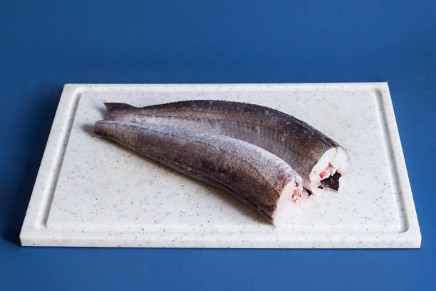 Two fresh pieces of headless hake on a white cutting board on a blue studio background.