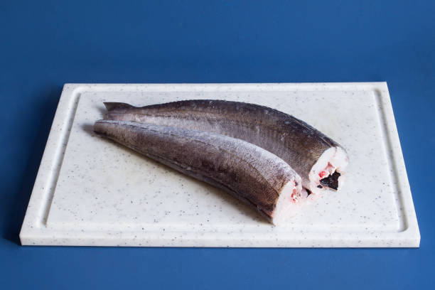 Two fresh pieces of headless hake on a white cutting board on a blue studio background. Two fresh pieces of headless hake on a white cutting board on a blue studio background. White fish and health. hake stock pictures, royalty-free photos & images