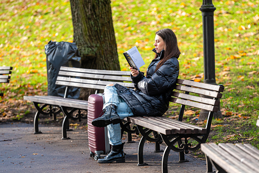 Riga, Latvia - November 4, 2022: Young woman reading a book and sitting on bench in autumn park. Autumn mood, leisure time concept.