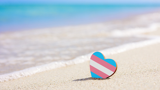 Transgender flag in the shape of a heart on the sandy beach against the background of the sea.