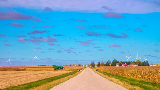 Hilly road across farmland in autumn, with corn (maize) field, farmstead, and wind turbines at right and a grain truck and turbines at left, southeast Illinois. Digital painting effect.