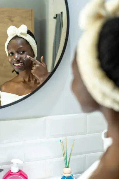 Young african woman doing daily cosmetic skin care routine while looking at herself in the mirror. Applying lotion on her cheek