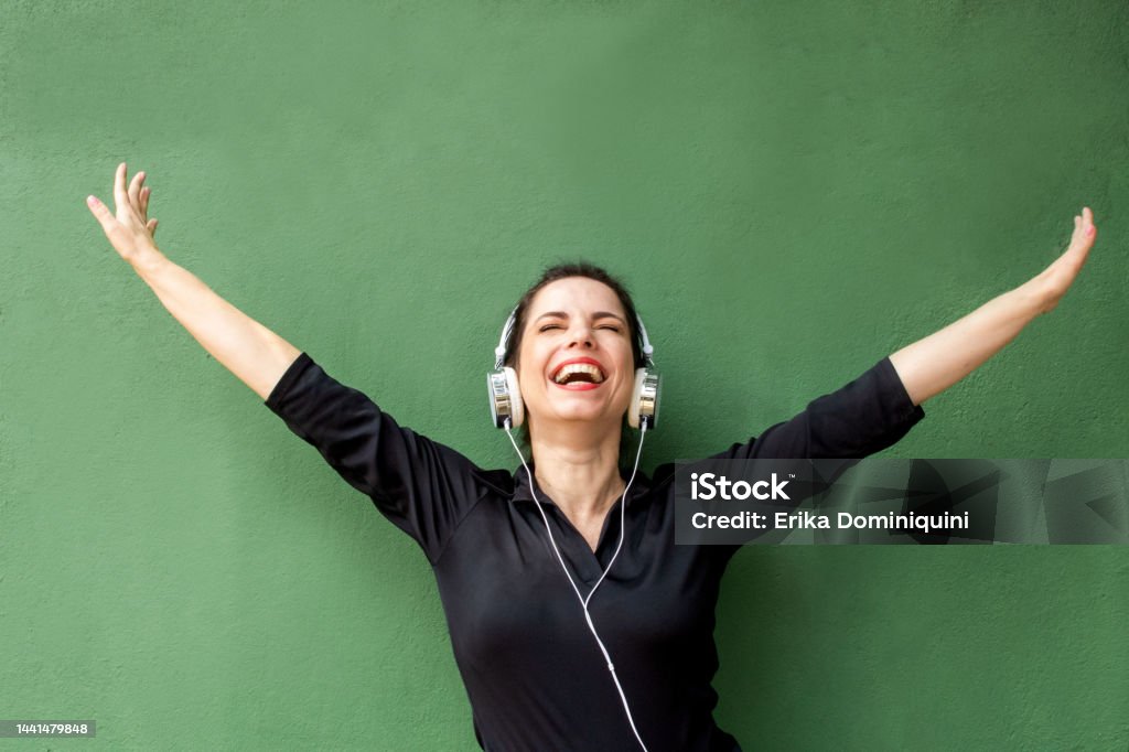 Girl in external photo Brunette girl in a black blouse and jeans, listening to music on the street, all happy near a green wall Emotion Stock Photo
