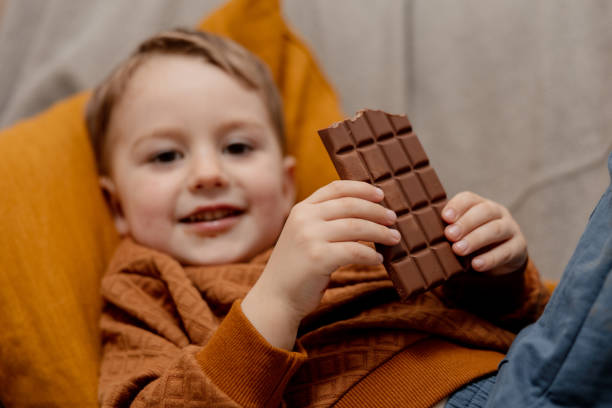 piccolo adorabile ragazzo seduto sul divano di casa e mangiando barretta di cioccolato. bambini e dolci, dolciumi di zucchero. i bambini si godono un delizioso dessert. bambino in età prescolare con abbigliamento casual. emozione positiva. - brown boy foto e immagini stock