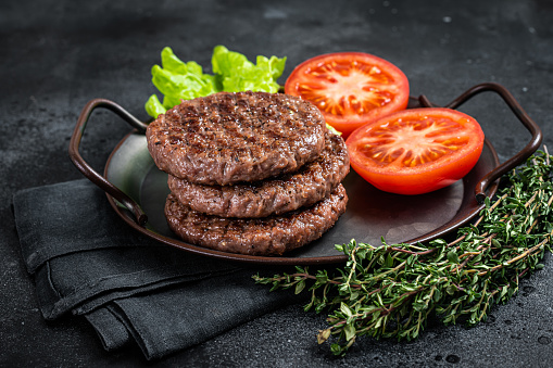 Tasty grilled burger beef patty with tomato, spices and lettuce in kitchen tray. Black background. Top view.