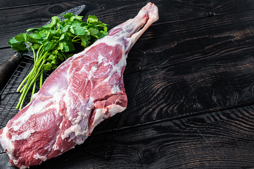 Fresh Raw whole lamb mutton leg thigh on kitchen table with herbs. Black Wooden background. Top view. Copy space.