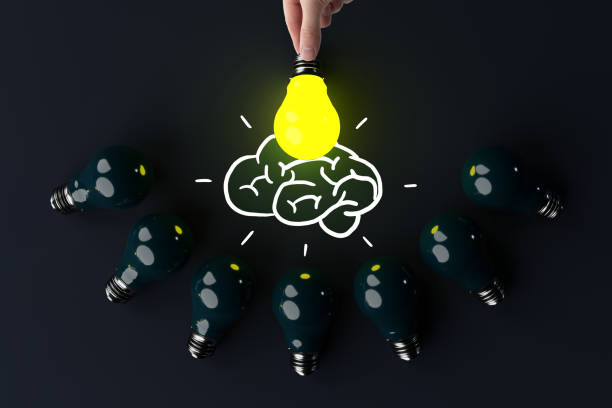 A hand holds a lit light bulb against the background of other extinguished light bulbs and a sketch of the brain.