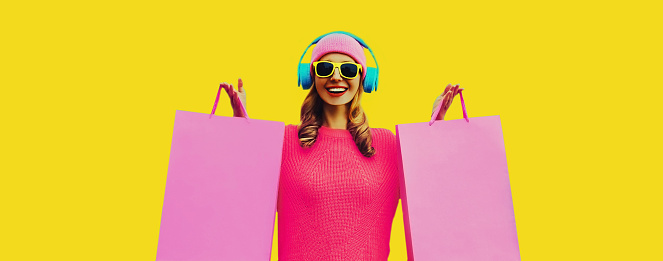 Portrait of stylish happy smiling young woman enjoying listening to music in headphones with colorful shopping bags posing wearing pink knitted sweater, hat on yellow background
