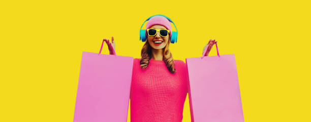 Portrait of stylish happy smiling young woman enjoying listening to music in headphones with colorful shopping bags posing wearing pink knitted sweater, hat on yellow background stock photo
