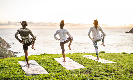 Rear view of group of three people standing on one leg and doing yoga on mat along the sea in morning