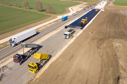 Aerial view of an asphalting paver machine and heavy road roller compacting freshly laid asphalt road surface of a new road.