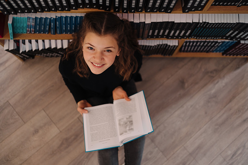 Teen girl among a pile of books. A young girl wearing glasses reads a book with shelves in the background. She is surrounded by stacks of books. Book day.