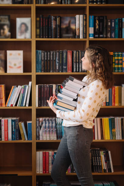 Beautiful girl is holding stack of books while standing among books in the bookshop stock photo