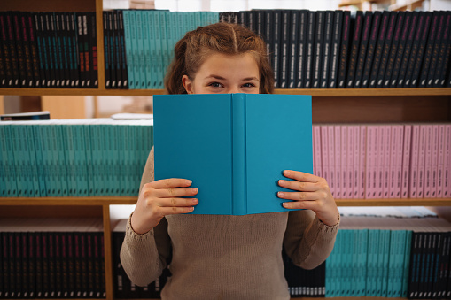 Portrait of a pretty young girl hiding behind an open blue book and looking away over bookshelf background