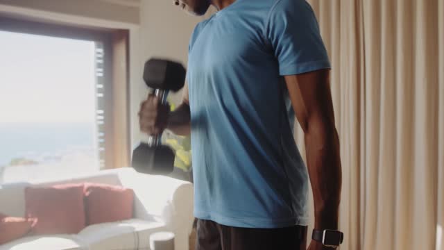 Front view of a Black African American young male exercising at home in his lounge lifting weights and dumbbells, bicep curls, keeping fit and healthy