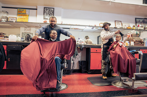 A Black woman, young child, teenager, and adult male receives a haircut and a line-up from a classic barbershop.