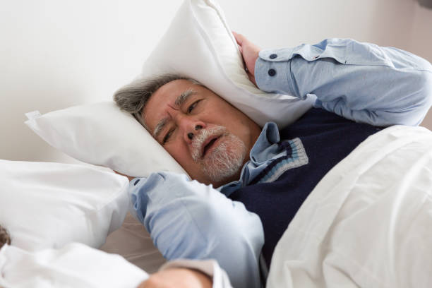 istock Elderly Asian man having trouble sleeping squeezes white pillow around ears for peace and quiet on bed in bedroom 1441552899