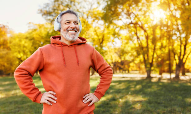 heureux homme mûr positif avec un large sourire au casque tout en faisant du sport dans le parc de la ville - adult jogging running motivation photos et images de collection