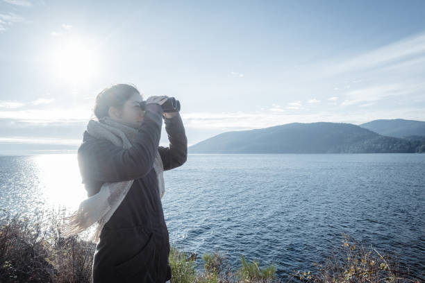 Multiracial young woman birdwatching from rocky viewpoint at Whytecliff Park, West Vancouver, British Columbia, Canada.  Bowen Island in the background.