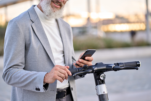 Old senior adult business man holding smartphone using electric bike rental digital phone mobile app renting scooter in city public eco transport mobile application standing in urban park. Closeup