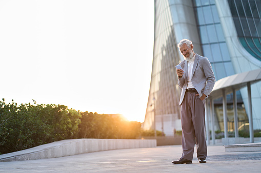 Happy older mature adult professional business man, smiling senior old businessman wearing suit holding smartphone using mobile cellphone standing outdoor in big city office downtown on sunset.