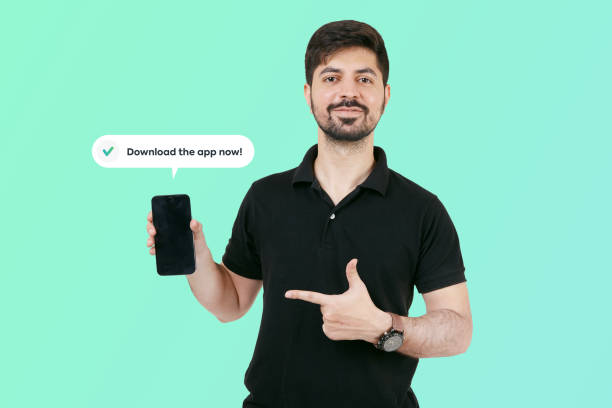 istock Happy Guy having smart phone with black screen in hand, pointing with forefinger to product. Guy holding mobile and asking to download the app now. Happy Indian guy showing mobile screen. 1441565012