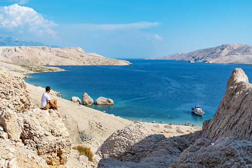 Person sitting on edge of cliff on rocky coastline near spectacular beach with rock formations in water on Beritnica beach on Pag island in Croatia.