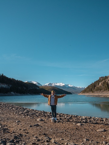 A vertical shot of a young Caucasian guy standing on the lake shore with his arms open