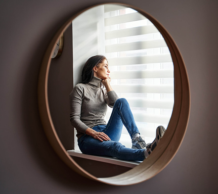 A young Latvian woman in thoughts sitting on a windowsill looking outside reflected on a round mirror