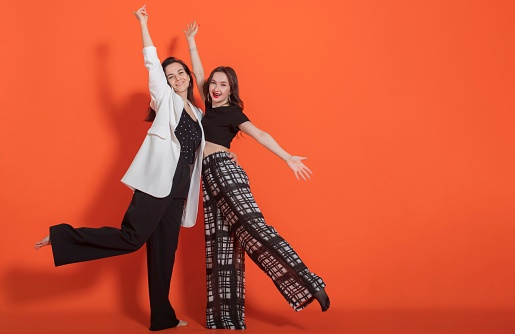 Two young brunette girls (family - mother and daughter), in casual clothes, dance on orange background, look at camera and laugh.