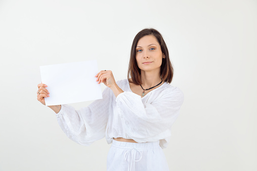 Portrait of young woman with short dark hair wearing white loose blouse, trousers, standing, holding blank sheet of paper, looking at camera on white background. Presentation, advertisement. Studio.