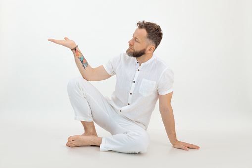 Portrait of tranquil sporty middle-aged bearded man with short grey dark hair wearing white shirt, trousers, sitting with bent knees on floor, raising hand with open palm on white background. Sport.