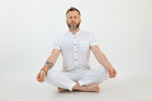 Portrait of middle-aged bearded man with grey dark hair wearing white shirt, trousers, sitting with crossed legs on floor, doing Sukhasana exercise with closed eyes on white background. Meditation.