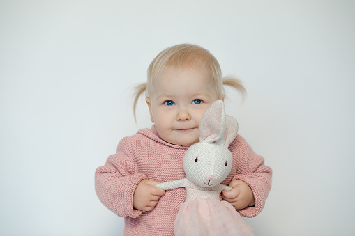 Cute baby girl play with stuffed animal, isolated on white. Smiling toddler gladly holding by hands teddy bunny. Blonde haired child in pink clothes with paschal rabbit. Easter. Holiday gift.