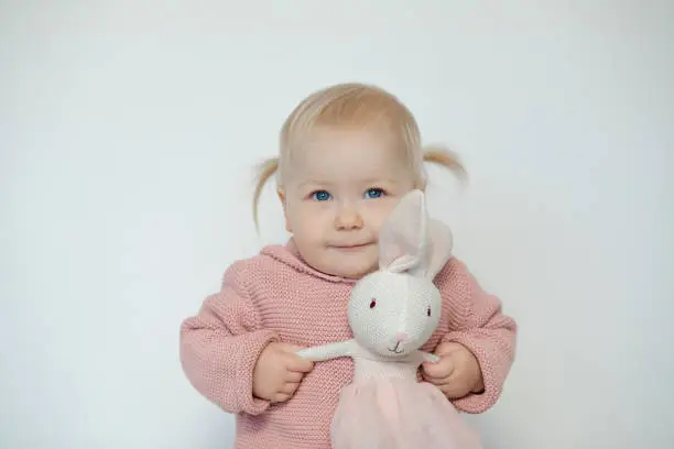 Photo of Cute baby girl play with stuffed animal, isolated on white. Smiling toddler gladly holding by hands teddy bunny. Blonde haired child in pink clothes with paschal rabbit. Easter. Holiday gift