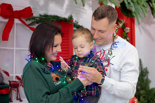 Family of three people hug together on christmas shooting location with red and green decorations. Joyful mother, father and little son hung with colored garland. New year, holiday, family look.