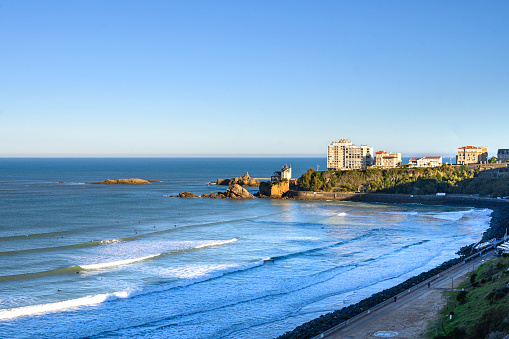 back view of biarritz beach on a sunny autumn morning. The sun hits the white buildings of the beach on the cliffs, below the beach the promenade in the shade, the waves break calmly against the sand of the beach