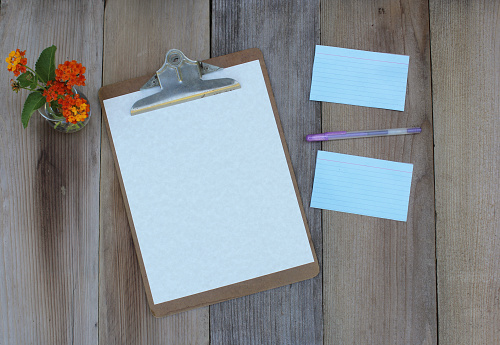 The flat lay of a clipboard with a blank sheet of paper on the wooden table with flowers