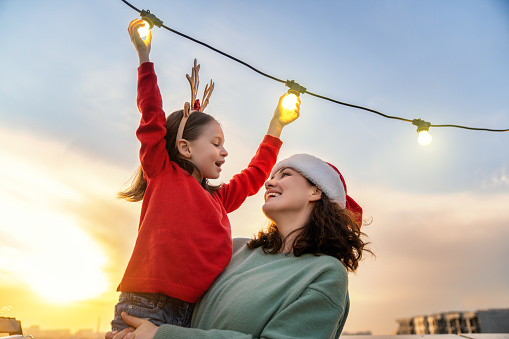 Merry Christmas and Happy Holidays! Happy mother and daughter are decorating home terrace or balcony with garlands.