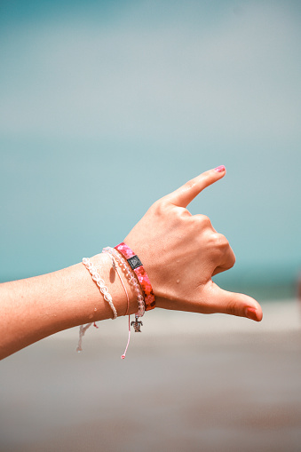 A vertical shot of a hand with bracelets doing a hang loose sign on a beach