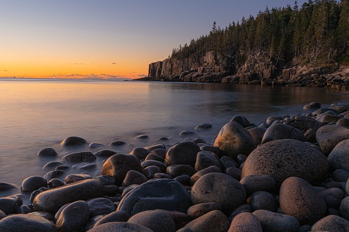 A beautiful scene of Boulder Beach in Acadia National Park, Maine with green hills and an orange sky