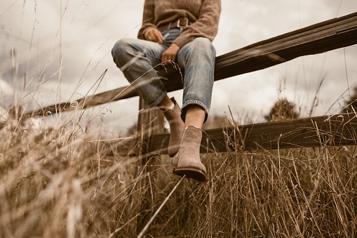 A close-up shot of boots of a woman sitting on a wooden fence in a rustic field - shoe advertisement
