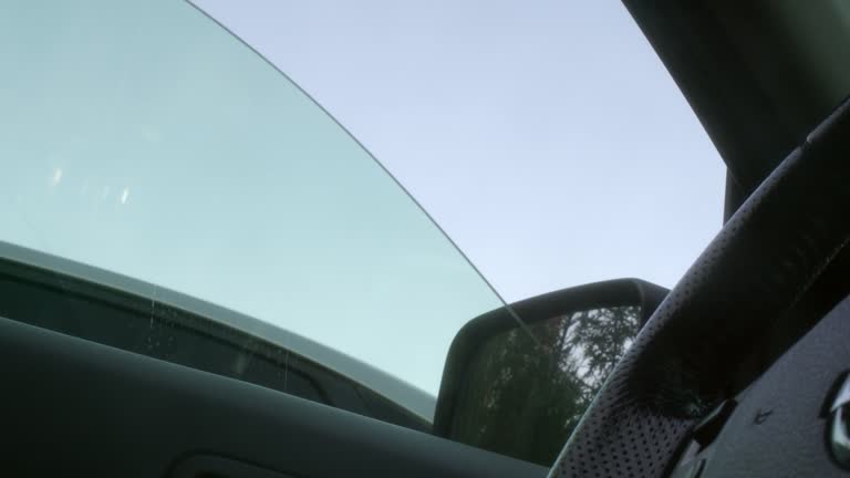 Close-up of closing and opening dirty car window covered in dust with blue sky on background. Driver's front side door automatic glass with leather steering wheel in foreground.