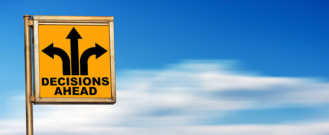 Closeup of a decisions ahead road sign (orange background and black arrows) against a blurred clear sky with clouds and copy space.