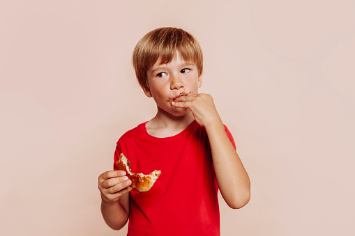 Portrait of a little cute blond kid eats chocolate bun, sucks fingers and holds bakery. Wears red t-shirt, has chocolate on face and hands and looks away. Studio shot isolated over beige background. Copy space.