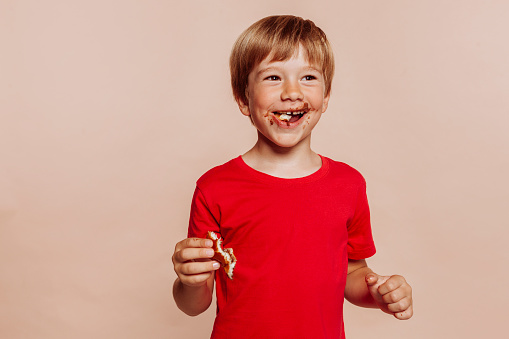 Portrait of a little cute blond cheerful kid eats chocolate bun and holds bakery. Wears red t-shirt, has chocolate on face, hands and looks away. Studio shot isolated over beige background. Copy space.