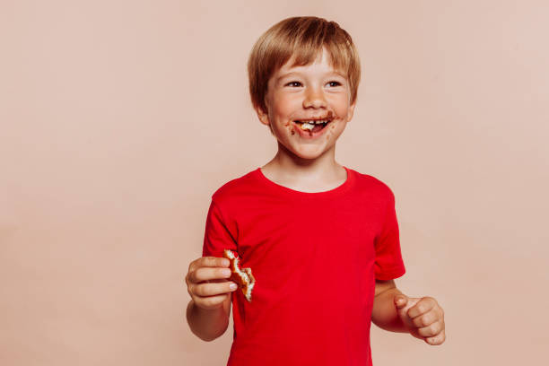 Little cute cheerful kid eats chocolate bun Portrait of a little cute blond cheerful kid eats chocolate bun and holds bakery. Wears red t-shirt, has chocolate on face, hands and looks away. Studio shot isolated over beige background. Copy space. candy in mouth stock pictures, royalty-free photos & images